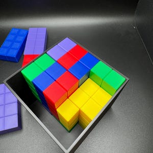 May include: A colorful cube puzzle with a black frame. The puzzle is composed of smaller blocks in various colors, including red, blue, green, yellow, and purple. Additional blocks are visible outside the frame. The background is black.
