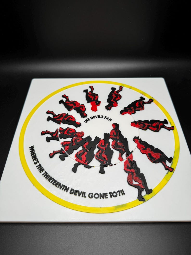 Op de afbeelding: Een witte vierkant met een gele cirkel en zwarte en rode figuren. De tekst "WHERE'S THE THIRTEENTH DEVIL GONE TO?!!" en "THE DEVIL'S FAN" zijn zichtbaar. De figuren zijn rond de cirkel gerangschikt.