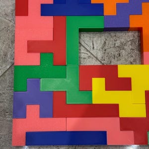 May include: A colourful puzzle made of plastic blocks in various shapes and colours, including red, green, blue, yellow, orange, and purple. The blocks are arranged in a pattern to form a square shape with a small opening in the centre.
