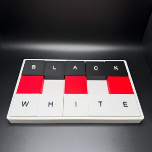 May include: A rectangular puzzle with the words "BLACK WHITE" spelled out in individual letter blocks. The blocks alternate between black, red, and white. The letters are in a sans-serif font.