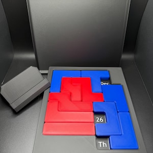 K&ouml;nnte beinhalten: Eine schwarze Puzzlebox mit einem blauen und roten Puzzle darin. Die Puzzleteile sind L-f&ouml;rmig und in einem Gittermuster angeordnet. Die Box hat den Text "Dec 26 Th" auf der Unterseite.