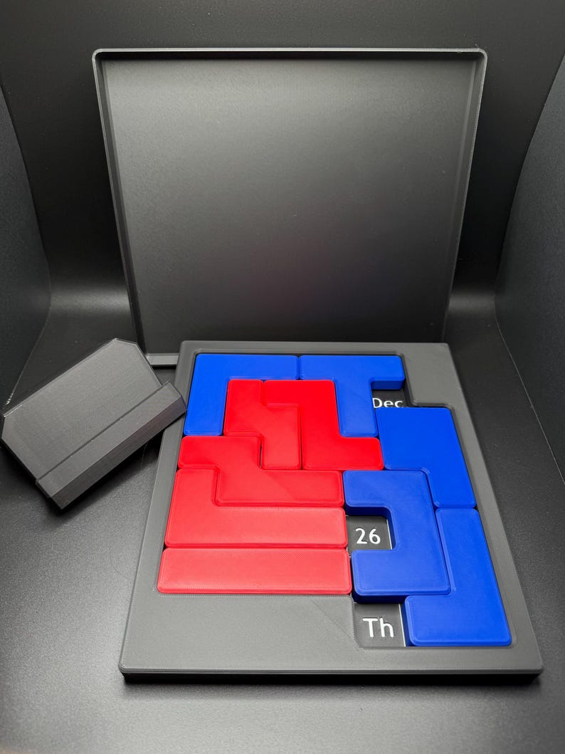 K&ouml;nnte beinhalten: Eine schwarze Puzzlebox mit einem blauen und roten Puzzle darin. Die Puzzleteile sind L-f&ouml;rmig und in einem Gittermuster angeordnet. Die Box hat den Text "Dec 26 Th" auf der Unterseite.