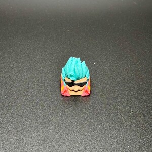 May include: A teal blue and pink 3D printed keycap with a cartoon face wearing sunglasses. The keycap has a spiky hairstyle.