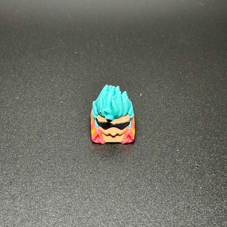May include: A teal blue and pink 3D printed keycap with a cartoon face wearing sunglasses. The keycap has a spiky hairstyle.