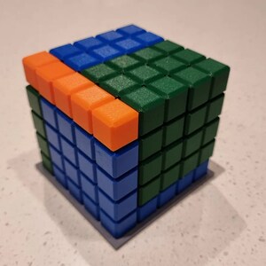 May include: A cube made of small square blocks in blue, green, and orange. The cube is made up of 64 blocks.