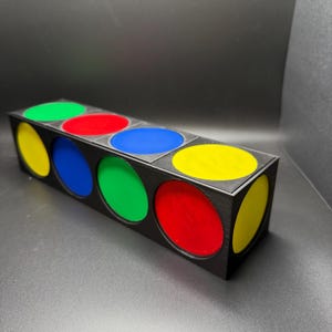 May include: A black rectangular box with six circular openings, each filled with a different colour: yellow, green, red, blue, green, and yellow. The box is on a dark surface.