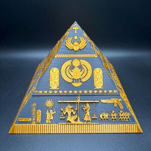 Puede incluir: Un objeto decorativo en forma de pir&aacute;mide, dorado y gris, con intrincadas tallas jerogl&iacute;ficas. La pir&aacute;mide presenta dise&ntilde;os intrincados de s&iacute;mbolos y figuras egipcias, incluyendo escarabajos, anj y faraones. La pir&aacute;mide es una detallada representaci&oacute;n del arte egipcio antiguo.
