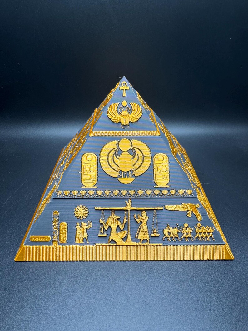Puede incluir: Un objeto decorativo en forma de pir&aacute;mide, dorado y gris, con intrincadas tallas jerogl&iacute;ficas. La pir&aacute;mide presenta dise&ntilde;os intrincados de s&iacute;mbolos y figuras egipcias, incluyendo escarabajos, anj y faraones. La pir&aacute;mide es una detallada representaci&oacute;n del arte egipcio antiguo.