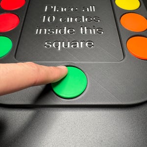 K&ouml;nnte beinhalten: Ein schwarzes Spielbrett mit zehn farbigen Kreisen und dem Text "Place all 10 circles inside this square". Ein Finger dr&uuml;ckt auf einen gr&uuml;nen Kreis. Die Kreise sind rot, blau, gelb und orange.