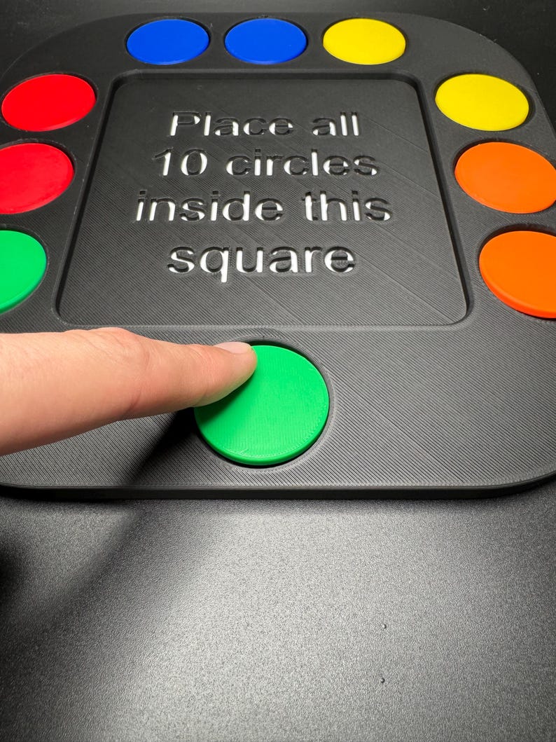 K&ouml;nnte beinhalten: Ein schwarzes Spielbrett mit zehn farbigen Kreisen und dem Text "Place all 10 circles inside this square". Ein Finger dr&uuml;ckt auf einen gr&uuml;nen Kreis. Die Kreise sind rot, blau, gelb und orange.