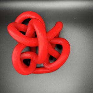 K&ouml;nnte beinhalten: Eine rote Plastikknotenskulptur. Der Knoten besteht aus einem einzigen St&uuml;ck Plastik, das verdreht und gedreht wird, um ein komplexes Design zu schaffen.
