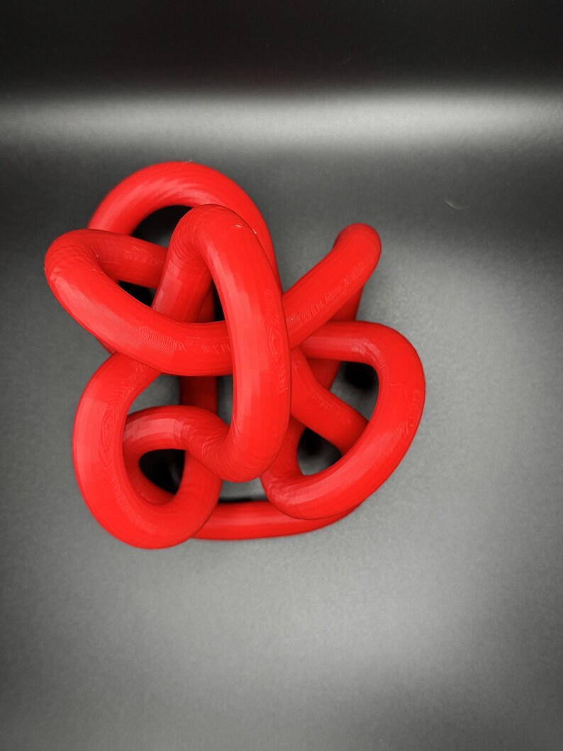 K&ouml;nnte beinhalten: Eine rote Plastikknotenskulptur. Der Knoten besteht aus einem einzigen St&uuml;ck Plastik, das verdreht und gedreht wird, um ein komplexes Design zu schaffen.