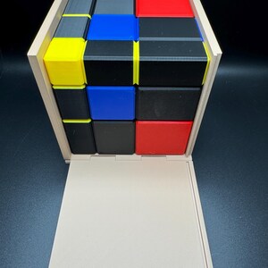 May include: A wooden box with a sliding lid, containing a 3x3 cube puzzle made of small blocks in red, blue, yellow, black, and grey.