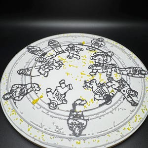 Puede incluir: Un plato de cer&aacute;mica blanco con un dise&ntilde;o gris y amarillo. El plato presenta un patr&oacute;n circular de astronautas en contornos negros. El borde del plato tiene un borde gris y est&aacute; salpicado de puntos amarillos.