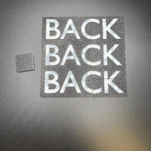 May include: A black square puzzle with the word "BACK" repeated three times in white block letters. A smaller square puzzle piece is next to the larger puzzle. The background is a dark gray, textured surface.