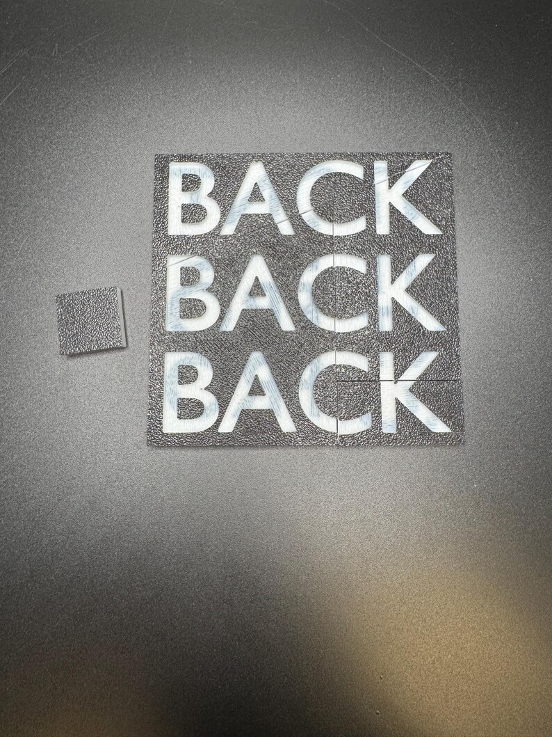 May include: A black square puzzle with the word "BACK" repeated three times in white block letters. A smaller square puzzle piece is next to the larger puzzle. The background is a dark gray, textured surface.
