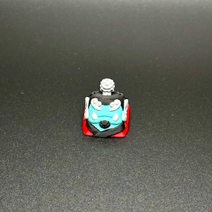 May include: A black and blue keycap with a white design and a red base. The keycap has a stylized face with a crown on top.