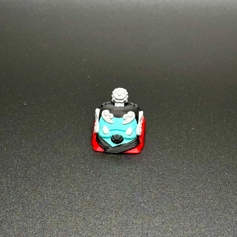 May include: A black and blue keycap with a white design and a red base. The keycap has a stylized face with a crown on top.