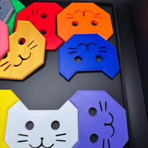 May include: A collection of colorful, cat-shaped puzzle pieces in various colors, including red, orange, blue, white, and purple. Each piece features a smiling cat face with eye and whisker details. The pieces are arranged in a black tray.