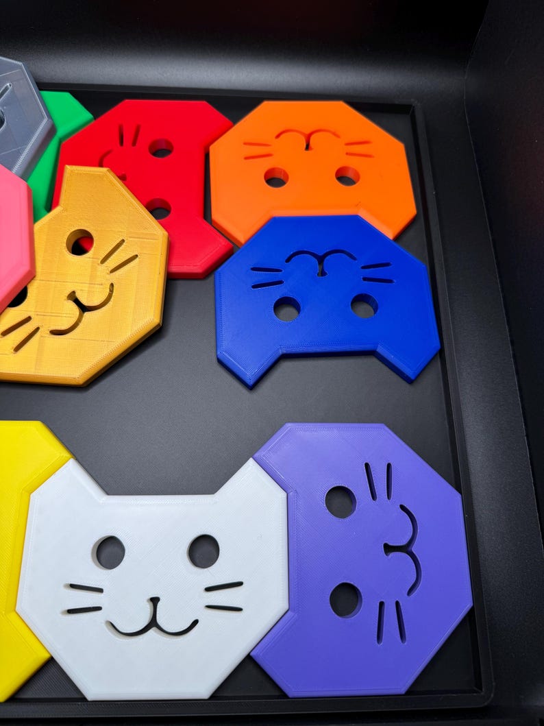 May include: A collection of colorful, cat-shaped puzzle pieces in various colors, including red, orange, blue, white, and purple. Each piece features a smiling cat face with eye and whisker details. The pieces are arranged in a black tray.