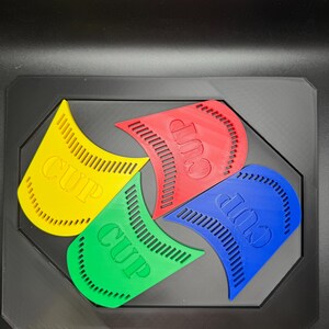 May include: Four brightly colored plastic cup holders in yellow, green, red, and blue, each with the word "CUP" printed on them. The holders are arranged in a black tray.