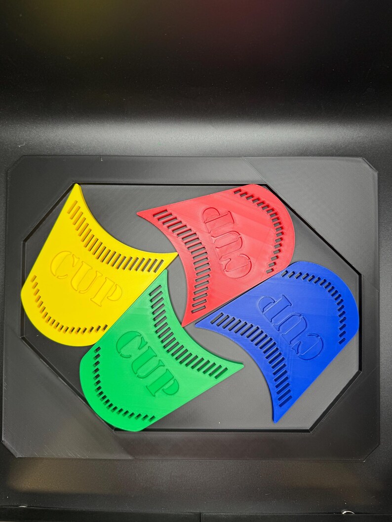 May include: Four brightly colored plastic cup holders in yellow, green, red, and blue, each with the word "CUP" printed on them. The holders are arranged in a black tray.
