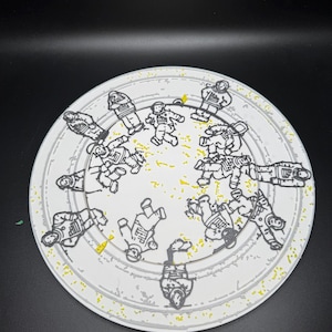 Puede incluir: Un plato decorativo redondo blanco con un patr&oacute;n repetido de figuras de astronautas negras. El plato tiene un borde gris y motas amarillas. Los astronautas se representan en varias poses, creando un dise&ntilde;o l&uacute;dico y tem&aacute;tico.