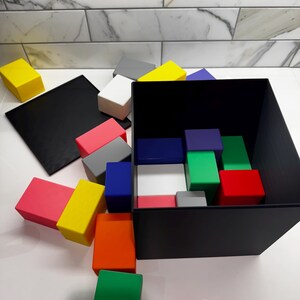 May include: A black box with a lid open, revealing a variety of colorful blocks inside. The blocks are red, green, blue, yellow, pink, gray, and white. There are also several blocks scattered around the box on a white surface.