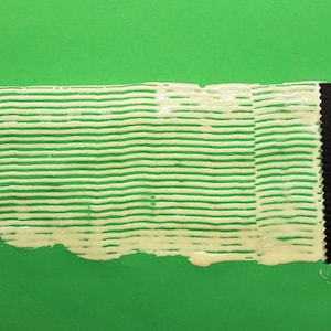 May include: A white, creamy substance is spread in parallel lines on a green surface using a black plastic tool. The substance is spread in a thin layer and has a slightly textured appearance.