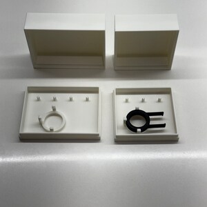 May include: Two white boxes with lids, one open and one closed. The open box contains a black ring with a white plastic insert. The closed box has a white plastic insert.