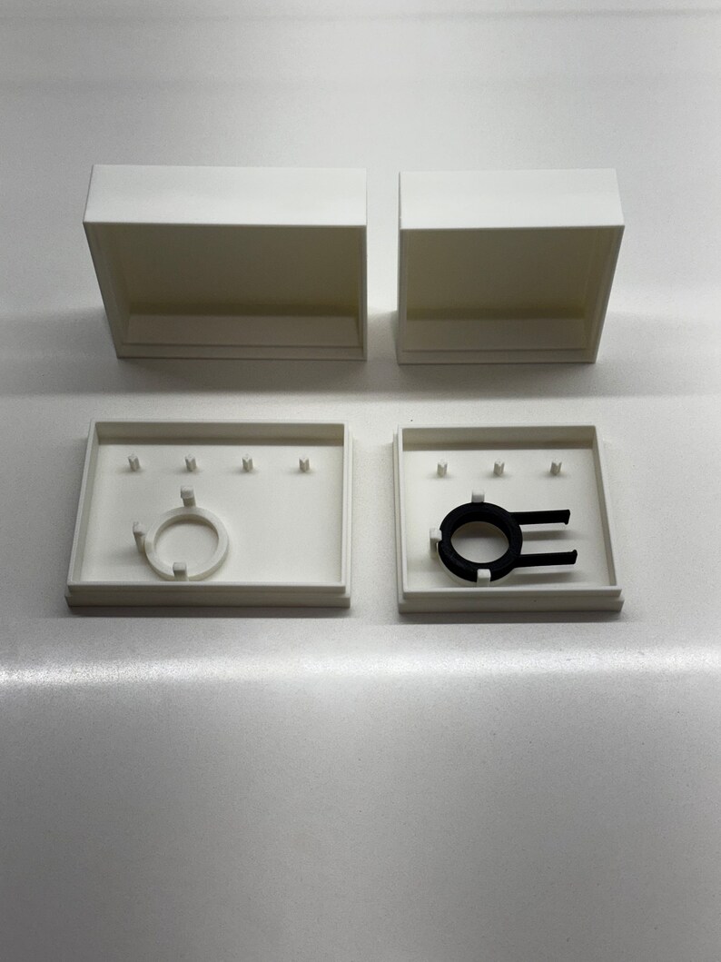May include: Two white boxes with lids, one open and one closed. The open box contains a black ring with a white plastic insert. The closed box has a white plastic insert.