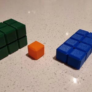 May include: Three 3D printed blocks, one green, one orange, and one blue. The green block is the largest and is made up of nine smaller cubes. The orange block is a single cube. The blue block is made up of six smaller cubes.