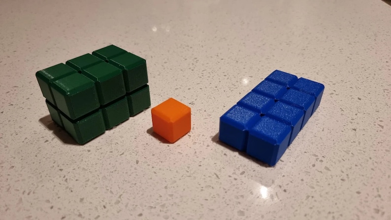 May include: Three 3D printed blocks, one green, one orange, and one blue. The green block is the largest and is made up of nine smaller cubes. The orange block is a single cube. The blue block is made up of six smaller cubes.