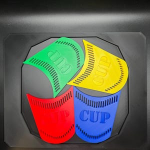 May include: A colorful, geometric design featuring four stylized cup shapes in green, yellow, red, and blue. Each cup has the word "CUP" printed on it. The design is set against a black background.