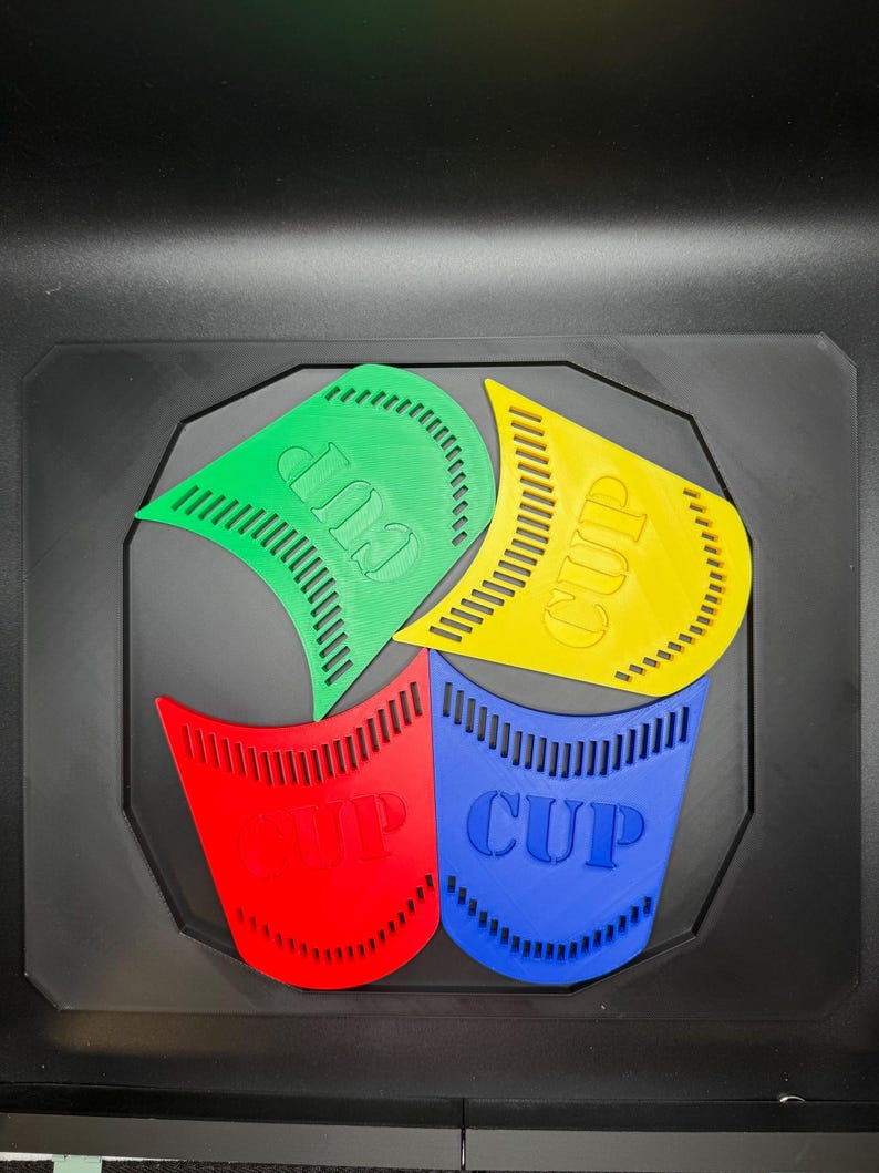 May include: A colorful, geometric design featuring four stylized cup shapes in green, yellow, red, and blue. Each cup has the word "CUP" printed on it. The design is set against a black background.