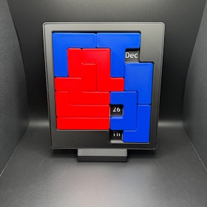 K&ouml;nnte beinhalten: Ein schwarzer Desktop-Kalender mit einem roten und blauen Puzzle-Design. Das Datum ist der 26. Dezember.