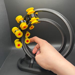 May include: A black, 3D-printed kinetic sculpture with yellow and red accents. The sculpture features a circular track with several yellow and red rollers. The base is black and the design is modern and geometric.