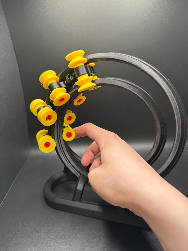 May include: A black, 3D-printed kinetic sculpture with yellow and red accents. The sculpture features a circular track with several yellow and red rollers. The base is black and the design is modern and geometric.