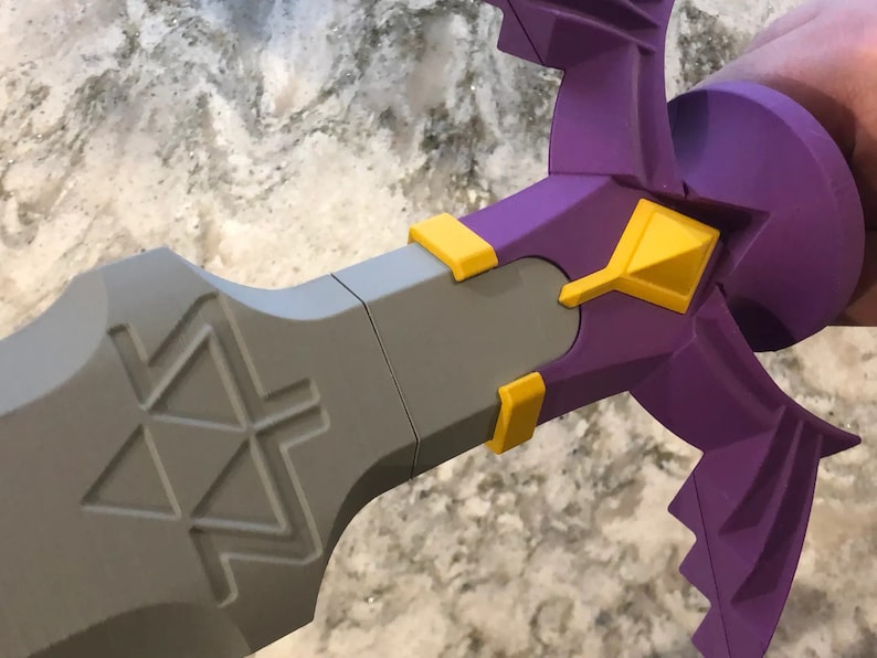 May include: A purple and yellow plastic sword with a triangular design on the blade. The sword is held by a hand.