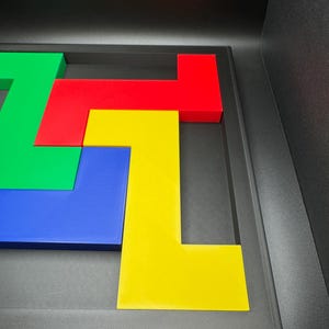 Op de afbeelding: Een kleurrijke puzzel met vier L-vormige stukken in groen, rood, blauw en geel, gerangschikt in een donkergrijs frame. De stukken zijn in elkaar verweven en cre&euml;ren een geometrisch ontwerp. Het frame heeft een getextureerd oppervlak.