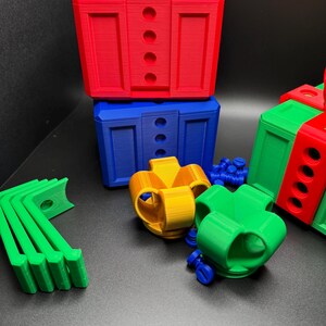 May include: A set of colorful 3D printed toy blocks in red, blue, green, and yellow. The blocks are shaped like presents and have holes in them. There are also some small green and blue plastic pieces.