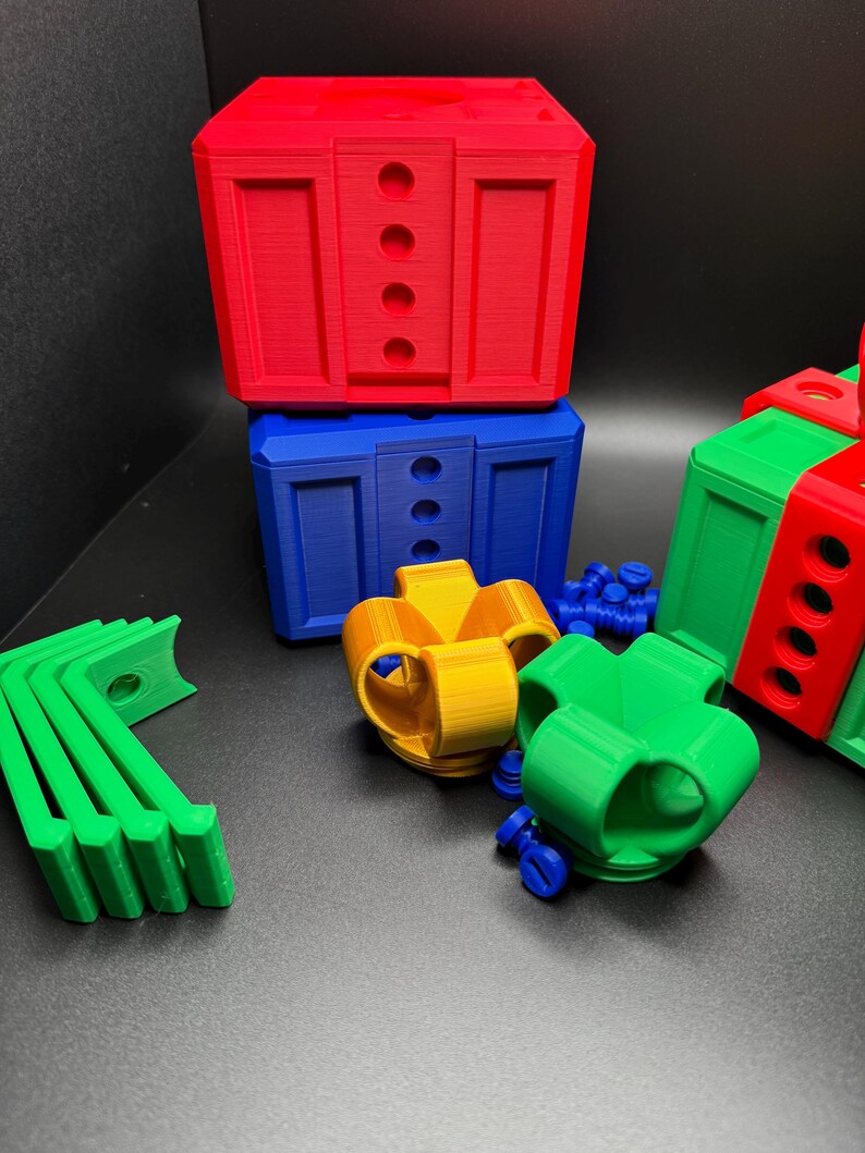 May include: A set of colorful 3D printed toy blocks in red, blue, green, and yellow. The blocks are shaped like presents and have holes in them. There are also some small green and blue plastic pieces.
