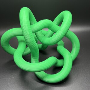 K&ouml;nnte beinhalten: Eine leuchtend gr&uuml;ne 3D-gedruckte Skulptur eines Knotens. Der Knoten besteht aus einem einzigen durchgehenden St&uuml;ck Plastik und ist als Dekorationsobjekt konzipiert.