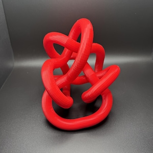 Può includere: Una scultura di nodo stampata in 3D di colore rosso brillante. Il nodo è realizzato in un unico pezzo continuo di plastica ed è progettato per essere un oggetto decorativo.