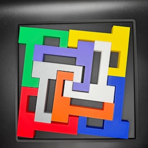 Op de afbeelding: Een kleurrijke puzzel met zeven stukken in een zwart frame. De stukken zijn T-vormig en zijn in een spiraalvorm gerangschikt. De kleuren van de stukken zijn rood, oranje, geel, groen, blauw, paars en zilver.