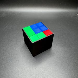 May include: A black cube puzzle with a colorful top. The top of the cube features a pattern of green, blue, and red squares. The cube is a classic brain teaser, perfect for puzzle enthusiasts.