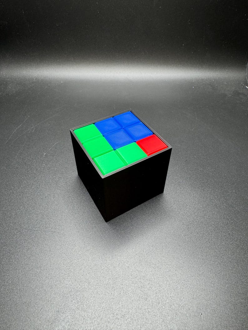 May include: A black cube puzzle with a colorful top. The top of the cube features a pattern of green, blue, and red squares. The cube is a classic brain teaser, perfect for puzzle enthusiasts.