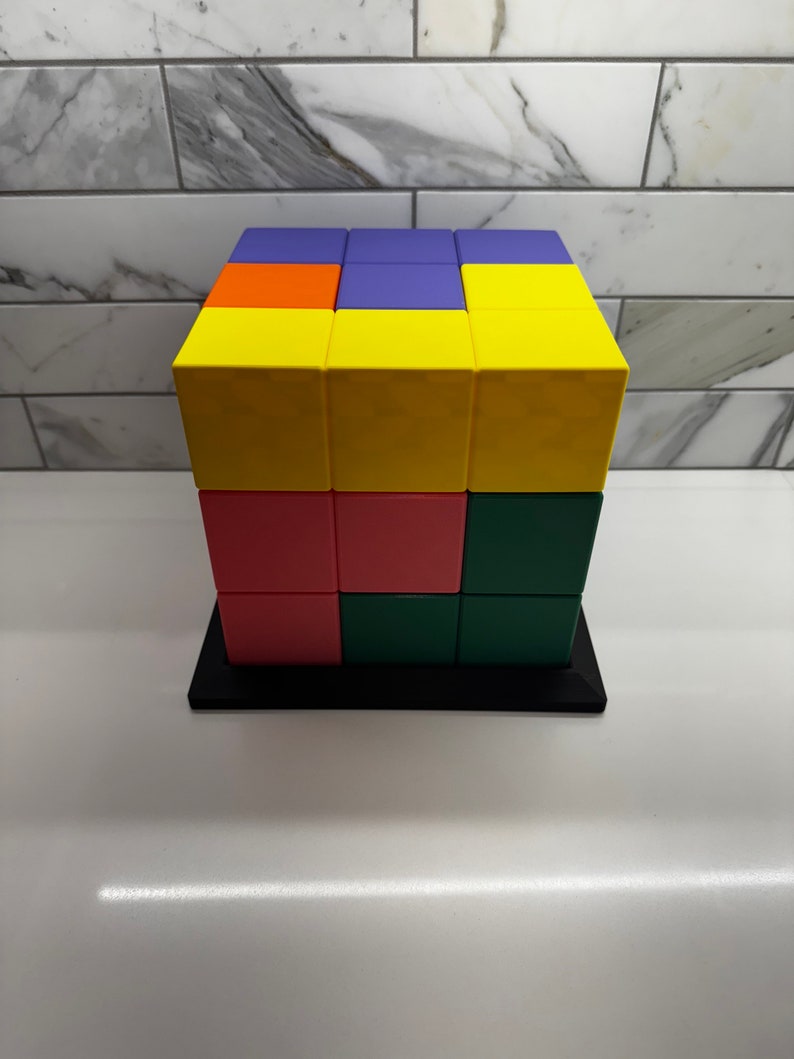 May include: A 3x3 cube made of colorful blocks. The top layer is yellow, orange, purple, and yellow. The middle layer is yellow, pink, green, and yellow. The bottom layer is pink, green, green, and yellow.