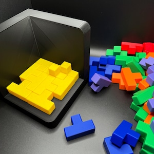 May include: A yellow Tetris-like puzzle piece set is assembled in a black frame. A pile of loose puzzle pieces in various colors, including blue, green, orange, and red, are scattered on a black surface.