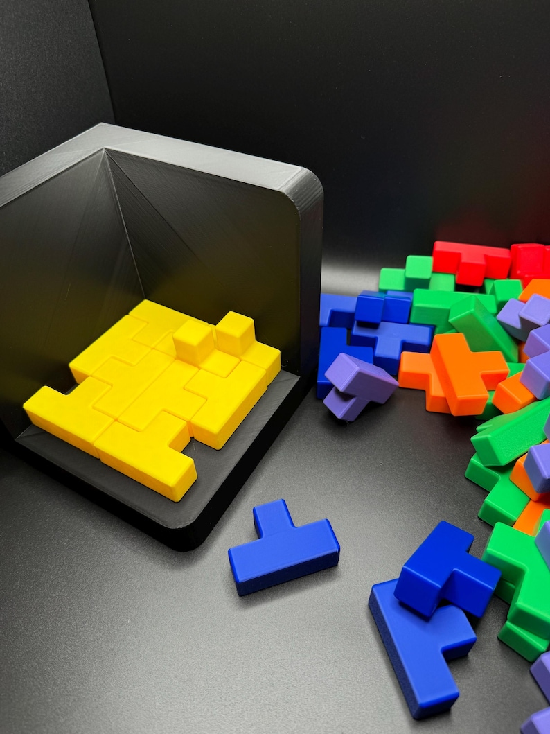 May include: A yellow Tetris-like puzzle piece set is assembled in a black frame. A pile of loose puzzle pieces in various colors, including blue, green, orange, and red, are scattered on a black surface.